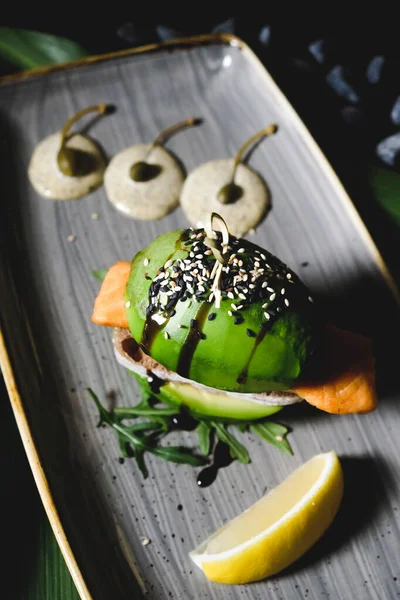 avocado burger with baked salmon and sesame seeds. avocado and salmon burger served in a restaurant on a gray plate decorated with a slice of lemon