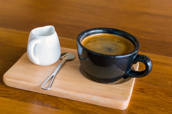 Taza de café azul oscuro con café americano, cuchara y una jarra de leche en un soporte de madera — Foto de Stock