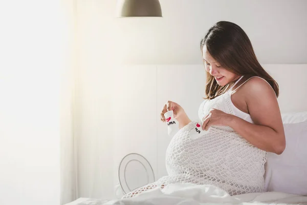Happy pregnant woman is resting at home on the bed and holding a little booties socks — Stock Photo, Image