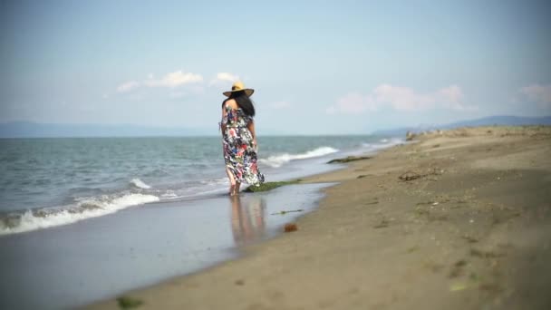 浪漫的女人穿着五颜六色的长裙 穿着风衣在海滩上散步 海浪正冲击着手里拿着拖鞋的女人的脚 一个人光着脚一个人 — 图库视频影像