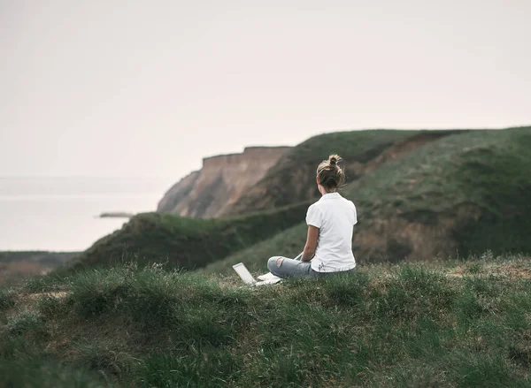 身穿白衬衫 金发的年轻女子 拿着笔记本电脑 坐在海滨附近的山上 望着前方 — 图库照片