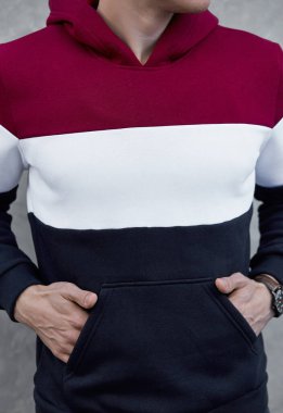 Gri arka planda duran kırmızı, beyaz ve mavi kapüşonlu genç adam.