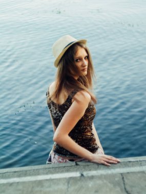 Gün batımında göl kenarında duran güzel boho kızı. Beyaz bohem elbiseli, rüzgarlı saçlı, yeşil yaprakların arasında, suyun yanında dinlenen hasır şapkalı çekici genç bir kadın. yaz tatili