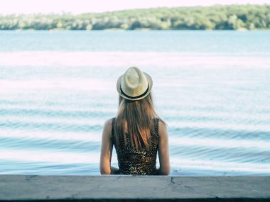 Gün batımında göl kenarında duran güzel boho kızı. Beyaz bohem elbiseli, rüzgarlı saçlı, yeşil yaprakların arasında, suyun yanında dinlenen hasır şapkalı çekici genç bir kadın. yaz tatili