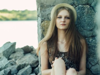 Gün batımında göl kenarında duran güzel boho kızı. Beyaz bohem elbiseli, rüzgarlı saçlı, yeşil yaprakların arasında, suyun yanında dinlenen hasır şapkalı çekici genç bir kadın. yaz tatili