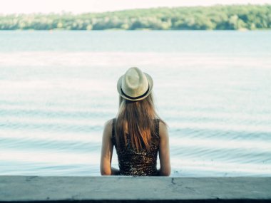 Gün batımında göl kenarında duran güzel boho kızı. Beyaz bohem elbiseli, rüzgarlı saçlı, yeşil yaprakların arasında, suyun yanında dinlenen hasır şapkalı çekici genç bir kadın. yaz tatili