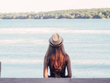 Gün batımında göl kenarında duran güzel boho kızı. Beyaz bohem elbiseli, rüzgarlı saçlı, yeşil yaprakların arasında, suyun yanında dinlenen hasır şapkalı çekici genç bir kadın. yaz tatili