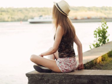 Gün batımında göl kenarında duran güzel boho kızı. Beyaz bohem elbiseli, rüzgarlı saçlı, yeşil yaprakların arasında, suyun yanında dinlenen hasır şapkalı çekici genç bir kadın. yaz tatili