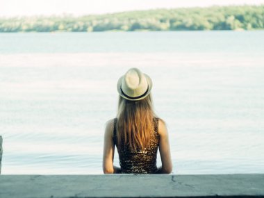 Gün batımında göl kenarında duran güzel boho kızı. Beyaz bohem elbiseli, rüzgarlı saçlı, yeşil yaprakların arasında, suyun yanında dinlenen hasır şapkalı çekici genç bir kadın. yaz tatili