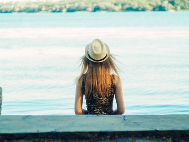 Gün batımında göl kenarında duran güzel boho kızı. Beyaz bohem elbiseli, rüzgarlı saçlı, yeşil yaprakların arasında, suyun yanında dinlenen hasır şapkalı çekici genç bir kadın. yaz tatili