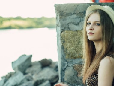 Gün batımında göl kenarında duran güzel boho kızı. Beyaz bohem elbiseli, rüzgarlı saçlı, yeşil yaprakların arasında, suyun yanında dinlenen hasır şapkalı çekici genç bir kadın. yaz tatili