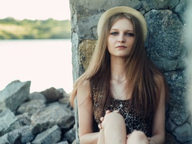 Gün batımında göl kenarında duran güzel boho kızı. Beyaz bohem elbiseli, rüzgarlı saçlı, yeşil yaprakların arasında, suyun yanında dinlenen hasır şapkalı çekici genç bir kadın. yaz tatili