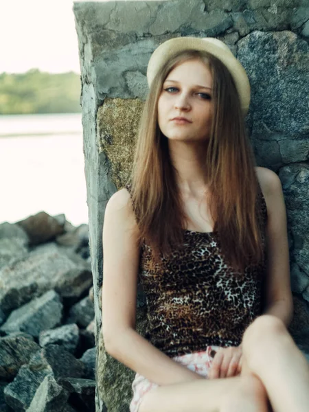 Gün batımında göl kenarında duran güzel boho kızı. Beyaz bohem elbiseli, rüzgarlı saçlı, yeşil yaprakların arasında, suyun yanında dinlenen hasır şapkalı çekici genç bir kadın. yaz tatili