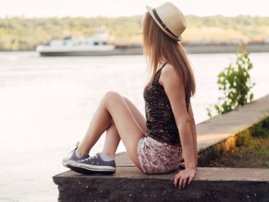 Gün batımında göl kenarında duran güzel boho kızı. Beyaz bohem elbiseli, rüzgarlı saçlı, yeşil yaprakların arasında, suyun yanında dinlenen hasır şapkalı çekici genç bir kadın. yaz tatili