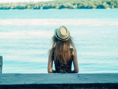 Gün batımında göl kenarında duran güzel boho kızı. Beyaz bohem elbiseli, rüzgarlı saçlı, yeşil yaprakların arasında, suyun yanında dinlenen hasır şapkalı çekici genç bir kadın. yaz tatili