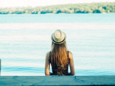 Gün batımında göl kenarında duran güzel boho kızı. Beyaz bohem elbiseli, rüzgarlı saçlı, yeşil yaprakların arasında, suyun yanında dinlenen hasır şapkalı çekici genç bir kadın. yaz tatili