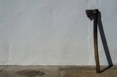 Old and dirty axe or ax resting on a white wall. Image of a rural place during a sunny day. Space in blank to add elements.