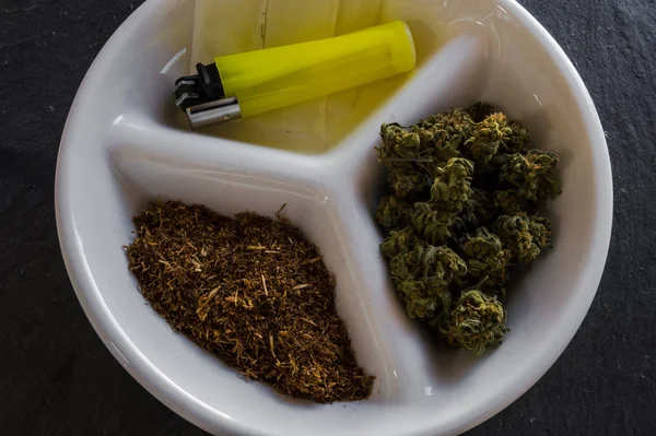 Modern and elegant plate with materials to roll a joint and smoke. Buds of marijuana, tobacco, lighter and rolling paper. Black rock background.
