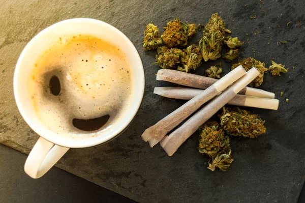 Lezzetli bir fincan kahve, yüksek kaliteli marihuana tomurcukları ve sigara içmeye hazır otlar. Siyah taşın arka planına sahip üst görünüm.