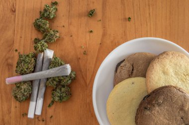 Pile of marijuana buds with rolled joints and a dish with homemade cookies of marijuana on wooden table. Breakfast of weed and cookies. clipart