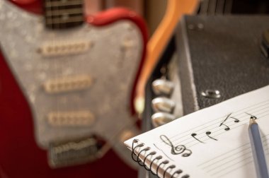 Composing music in a staff notebook with guitar and bass in the background. Musician writing in the music studio. Composing in treble clef. clipart