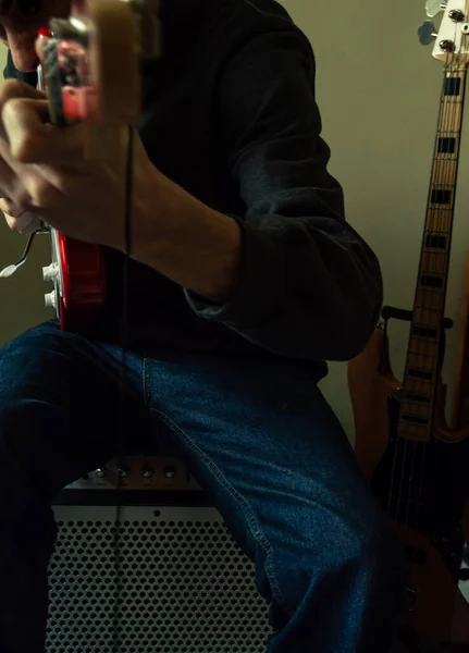 Young man sitting on a big speaker and playing a red electric guitar with a bass in the background. Playing electric guitar in aggressive mode.