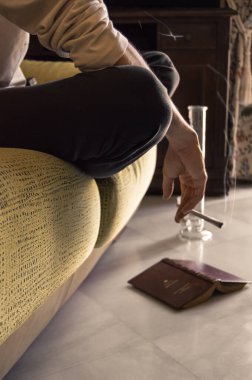 Kanepede oturan bir adam bir kitap okuduktan sonra esrar içer. Evde marihuana içmek, yerde kitap seyretmek ve arka planda büyük bir nargile..