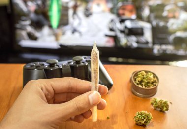 Video oyunları ve esrar. Elin büyük bir marihuana sigarası gösteriyor. Oyun denetleyicisi, marihuana tomurcukları, öğütücü ve video oyunu ile TV ile arka plan.