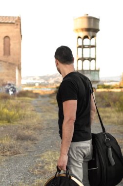 Man with casual clothes and luggage lost in the outskirts of the city at sunset. Exploring countryside and abandoned buildings in the urban suburbs. clipart