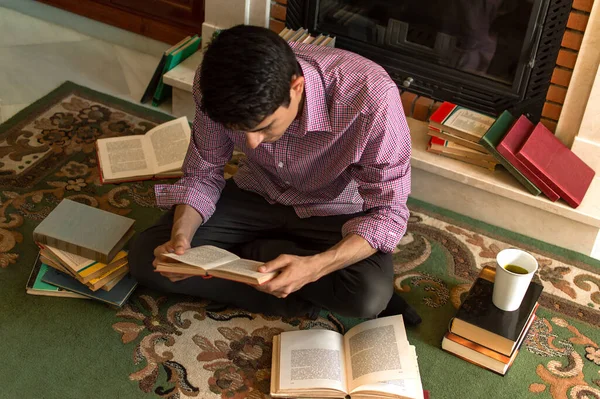Untidy young man reads a book sitting on a carpet full of books. Smart man reads concentrated at home surrounded by books in a classic living room.