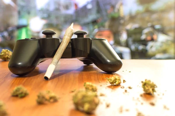 Video oyunları ve esrar. Oyun kumandası, büyük esrar ve marihuana tomurcukları ile ahşap masa. Video oyunu ile televizyonun bulanık arkaplanı.
