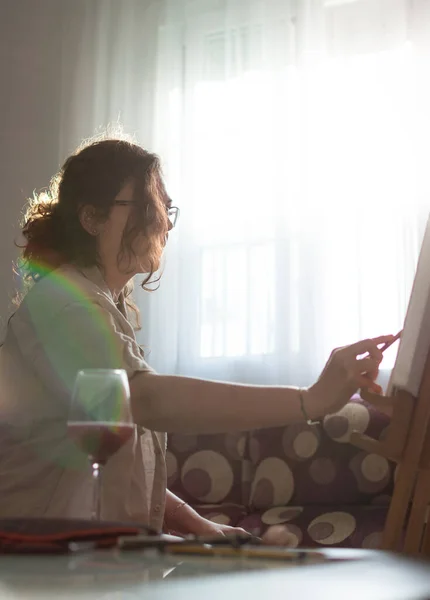 Woman with casual clothes painting a picture with crayons while having a glass of wine. Drawing at home in front of a bright window with easel.