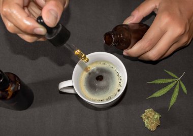 Marihuana 'nın doğal çaresi olarak kahve fincanına CBD tentürü dökmek. Marihuana yaprağıyla dolu bir masada marihuana yağı kullanan kişi..