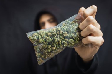 Siyah arka planda kafası önde büyük bir torba ot tutan kapüşonlu bir adam. Uyuşturucu ticareti kavramı, marihuana alıp satmak.