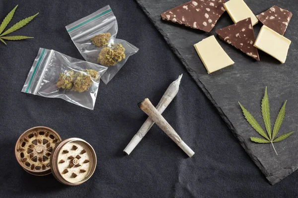 Esrar ve tatlılar: Esrar, marihuana yaprakları, plastik torba ve öğütücüdeki tomurcuklar ve siyah dokuma arka planındaki farklı çikolata türleri.