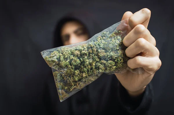 Siyah arka planda kafası önde büyük bir torba ot tutan kapüşonlu bir adam. Uyuşturucu ticareti kavramı, marihuana alıp satmak.