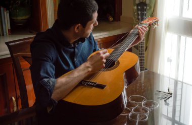 Genç adam parlak bir pencerenin yanında gitar tellerini değiştiriyor. Yeni teller ve aletlerle ahşap masada klasik bir gitarı tamir etmek.
