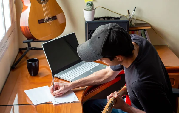 Man composing music and playing electric guitar using a laptop at home. Young musician in his room practicing guitar. Concept of learn music online.