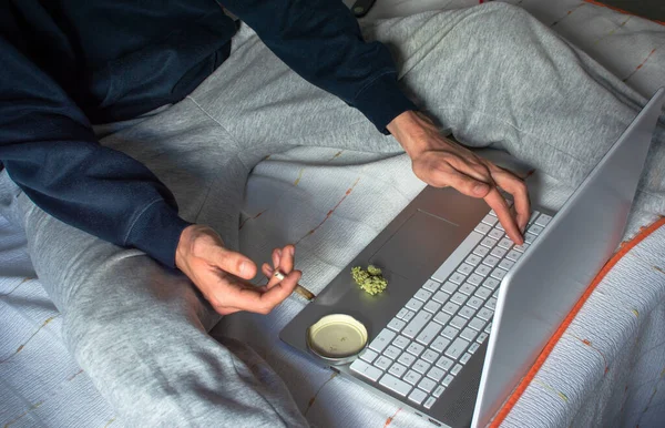 Top view of person sitting on a bed smoking marijuana joint while using a laptop at home. Cannabis and technology concept.