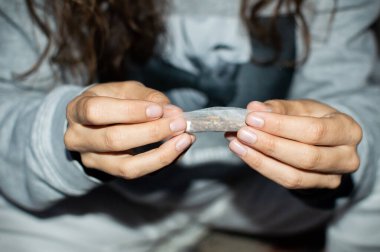 Young woman in tracksuit rolling a marijuana joint in the street at night. Details of hands rolling cannabis cigarette. clipart