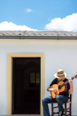 Hasır şapkalı genç adam güneşli bir günde kırsal kesimdeki beyaz evinin önünde gitar çalıyor..