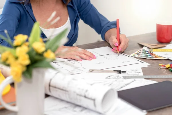 An architect sitting at a desk working on plans. There is a color swatch and red mug nearby and yellow flowers blurred in the foreground. 