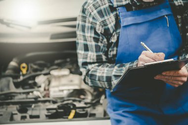 Araba tamircisi bir araba motorunu kontrol ediyor ve panoya yazı yazıyor.