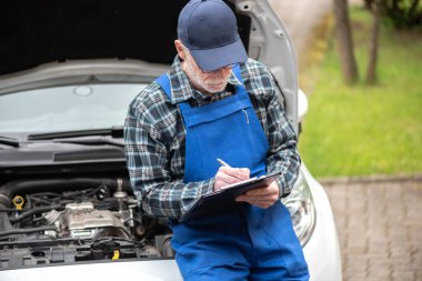 Araba tamircisi bir araba motorunu kontrol ediyor ve panoya yazı yazıyor.