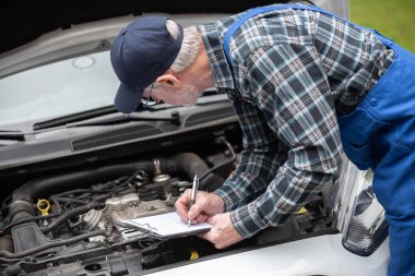 Araba tamircisi bir araba motorunu kontrol ediyor ve panoya yazı yazıyor.