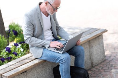 Olgun bir iş adamı dışarıda bir bankta oturuyor ve dizüstü bilgisayar kullanıyor. 