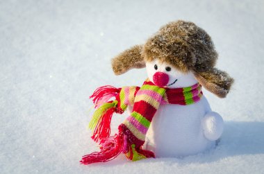 Kulaklıklı bir şapkalı oyuncak kardan adam ve güneş ışığıyla aydınlatılmış kar üzerinde parlak bir eşarp.