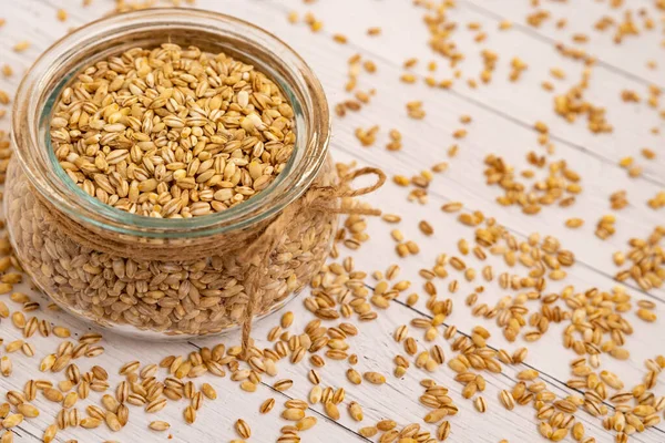 A close-up of pearl barley in a glass jar with a jute cord and scattered on a white wooden table.