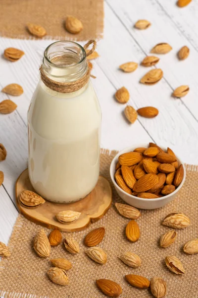 Almond milk in a glass bottle close-up, almonds in their shells and peeled in a glass white bowl and scattered on a jute napkin on a light table.
