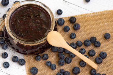 Böğürtlen reçelli cam kavanozun yakın çekimi ve beyaz ahşap bir masada ahşap bir kaşık, jüt peçeteye dağılmış dutlar..